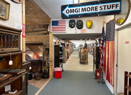 A hallway in an antique store with various items, an American flag, and a sign reading "OMG! MORE STUFF."