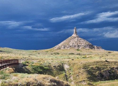 A dramatic landscape featuring a tall rock formation under a dark, cloudy sky, with a wooden bridge in the foreground.