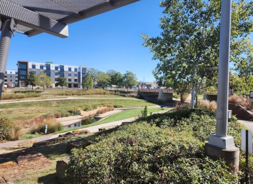 A sunny park scene with green grass, trees, and a bridge in the background, surrounded by modern buildings.