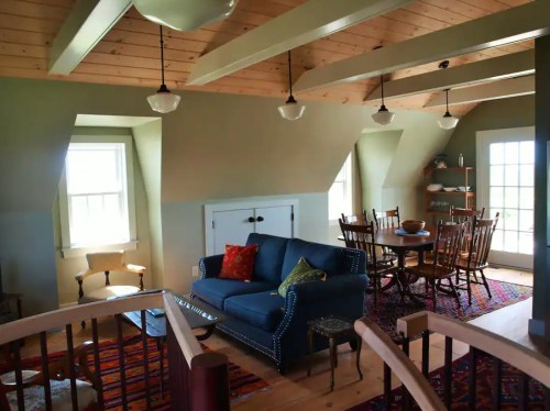 Cozy living space with a blue sofa, wooden dining table, and large windows, featuring warm wood accents and colorful rugs.