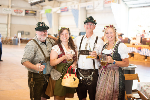 Four people in traditional Bavarian attire, smiling and holding drinks, inside a festive indoor venue.