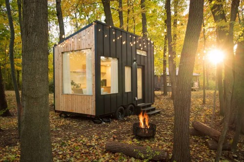 A modern tiny house with large windows, surrounded by trees and autumn foliage, with a fire pit in the foreground.