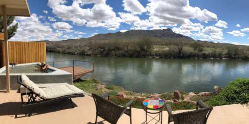 A serene riverside view with mountains in the background, featuring lounge chairs and a colorful table.