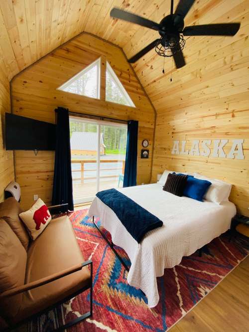 Cozy cabin interior with a bed, sofa, and large windows, featuring wooden walls and an "ALASKA" sign.