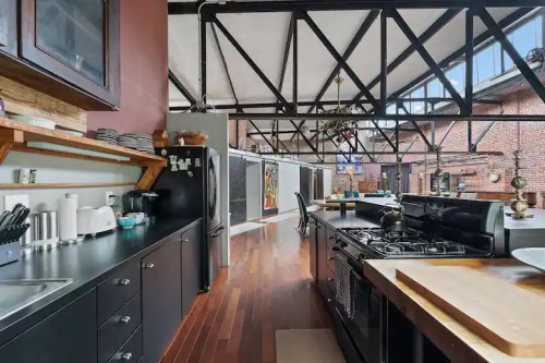 Modern kitchen with dark cabinetry, stainless steel appliances, and wooden floors, featuring an open layout and industrial beams.