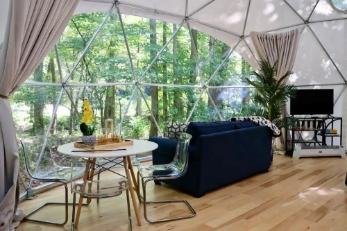Cozy interior of a geodesic dome with a sofa, dining table, and large windows overlooking a forested area.