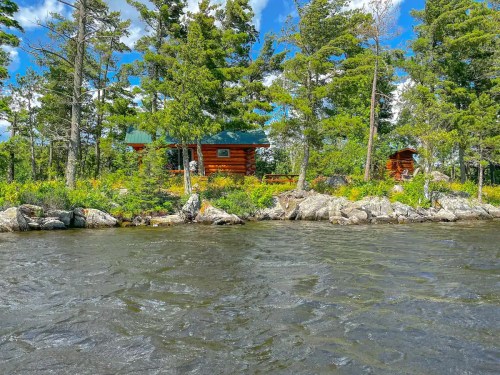 A rustic log cabin surrounded by trees on a rocky shoreline, with a calm lake in the foreground and blue skies above.