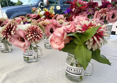 A table with mason jars filled with colorful flowers, labeled "Iron Hill," set against a backdrop of a blue vehicle.