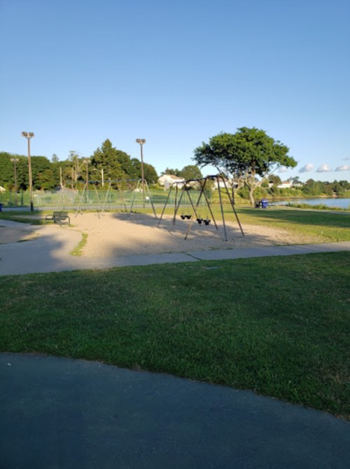 A playground with empty swings on a sandy area, surrounded by grass and trees under a clear blue sky.