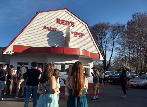 A group of people in line outside Red's Dairy Freeze on a sunny day, with cars parked nearby.