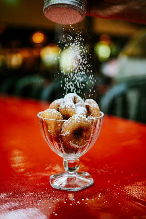 A glass bowl filled with donuts being dusted with powdered sugar on a red tablecloth.