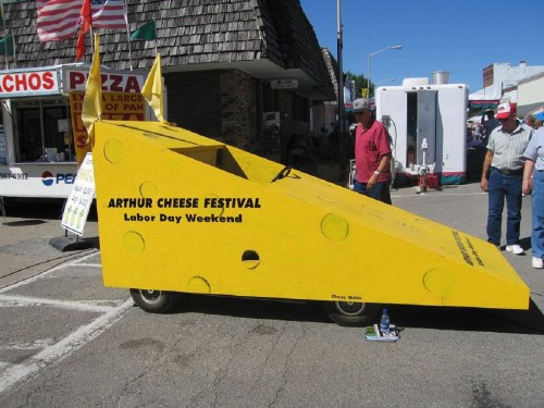 A large, yellow cheese-shaped vehicle at the Arthur Cheese Festival, with people nearby and food stalls in the background.