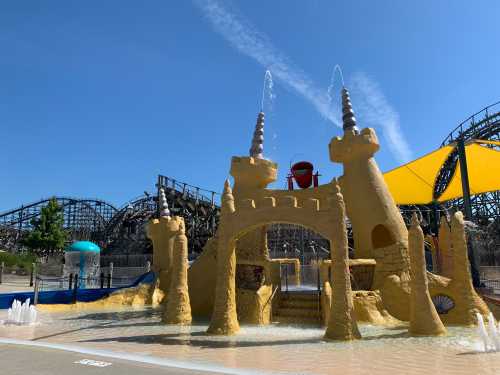 A colorful water play area with castle structures, fountains, and a roller coaster in the background under a clear blue sky.