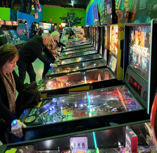 A group of people playing pinball machines in a colorful arcade filled with various games.