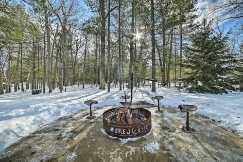 A snowy forest scene featuring a fire pit surrounded by metal benches and trees, with sunlight filtering through the branches.
