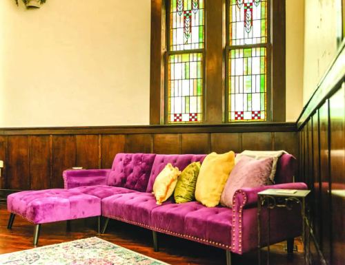 A purple sectional sofa with colorful pillows sits against a wooden wall, near a stained glass window.