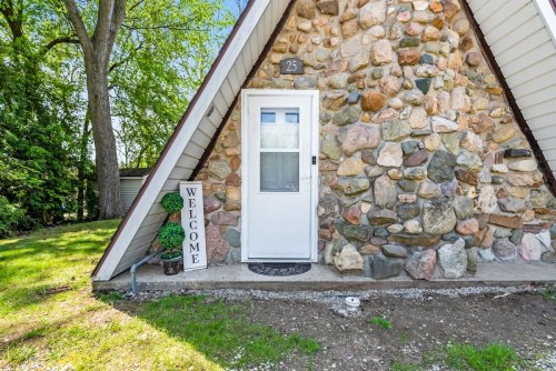 A stone-faced A-frame house entrance with a white door and a "Welcome" sign beside it, surrounded by greenery.