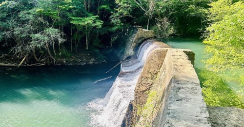A serene waterfall cascading over a stone dam, surrounded by lush green trees and clear blue water.