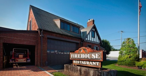 A brick building with a sloped roof and a sign reading "Firehouse Restaurant" in front, featuring a vintage fire truck inside.