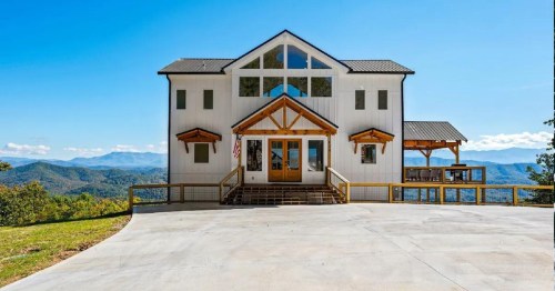 A modern white house with a wooden porch, set against a backdrop of mountains and a clear blue sky.