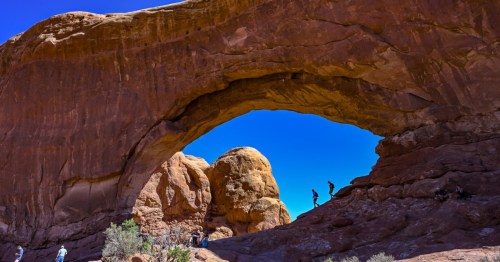 A natural rock arch with people exploring the area, surrounded by red rock formations and a clear blue sky.