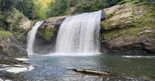 A serene waterfall cascades into a calm pool, surrounded by lush greenery and rocky cliffs.