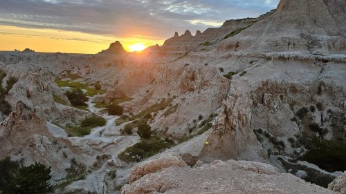 Sunset over rugged, eroded rock formations in a canyon, with soft clouds in the sky and greenery below.