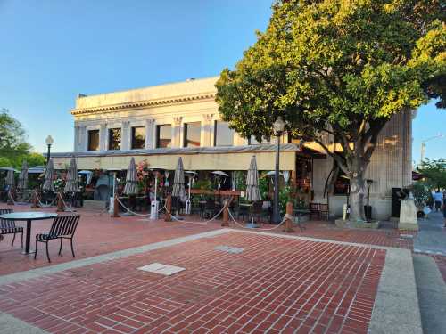 A charming restaurant with outdoor seating, surrounded by greenery and flowers, set against a clear blue sky.