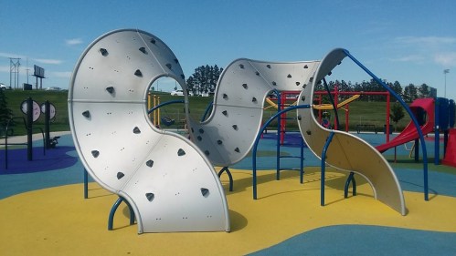 A modern playground structure with wavy climbing walls on a colorful rubber surface under a clear blue sky.
