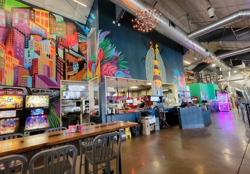 A vibrant restaurant interior featuring colorful murals, arcade games, and a bustling kitchen area.
