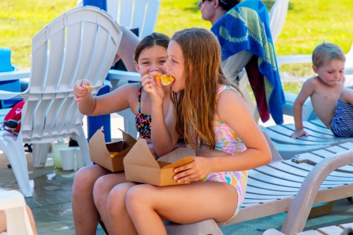 Two girls in swimsuits sit on lounge chairs, enjoying food from takeout boxes by a pool, with others in the background.