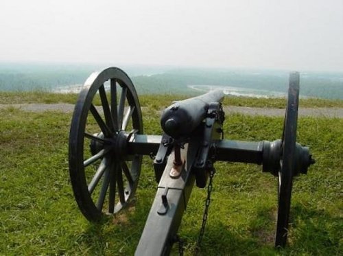A historic cannon on a grassy hill, overlooking a scenic landscape with trees and a river in the distance.