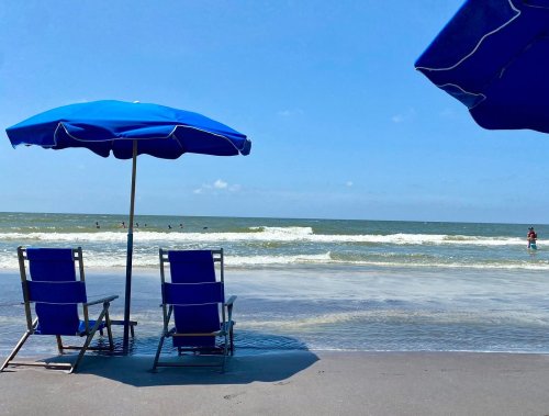 Two blue beach umbrellas shade empty chairs on a sandy shore, with gentle waves and people swimming in the background.