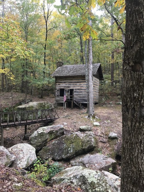 A rustic log cabin surrounded by trees and rocks, with an American flag hanging by the door.