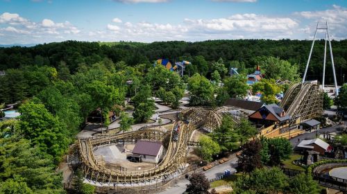 Aerial view of a wooden roller coaster surrounded by trees and amusement park attractions on a sunny day.