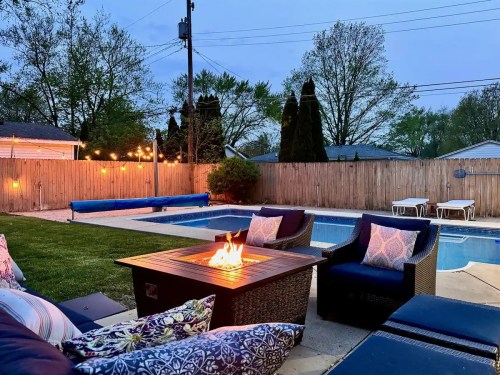 Cozy backyard with a fire pit, pool, and seating area, surrounded by trees and evening lights.