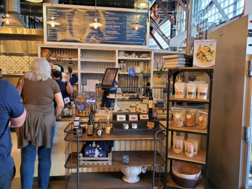 A busy café counter with a menu board, customers ordering, and shelves displaying various food products.