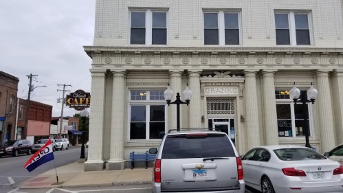 Historic building with columns, featuring a café sign, parked cars, and a flag in front.