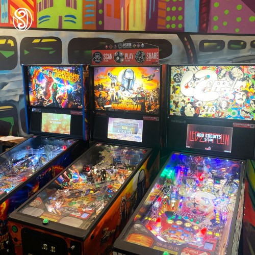 Three colorful pinball machines with vibrant artwork in a lively arcade setting.