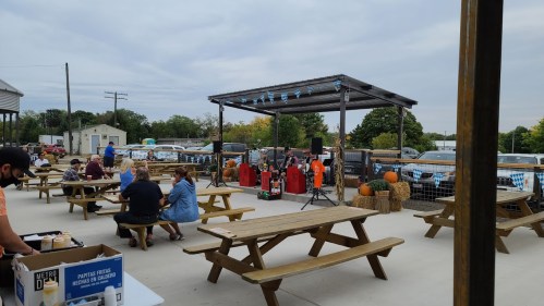 Outdoor venue with picnic tables, a stage for live music, and autumn decorations like pumpkins. People are enjoying the event.