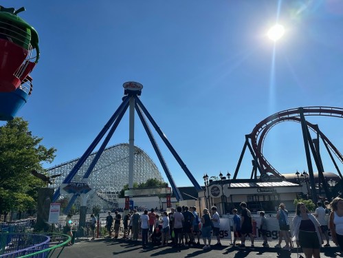 A sunny amusement park scene with a roller coaster, a ride, and a crowd of people waiting in line.