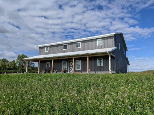 A modern gray farmhouse with a metal roof, surrounded by green grass and under a partly cloudy sky.