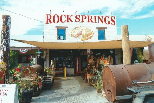 A rustic storefront labeled "Rock Springs," featuring outdoor decor, plants, and a large metal grill in front.