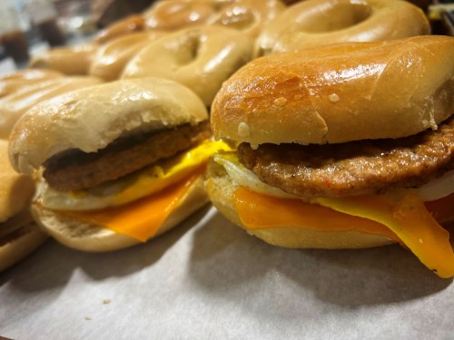 Close-up of a sandwich made with a glazed donut, sausage patty, and melted cheese, surrounded by more donuts.