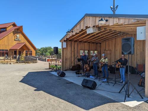 A band performs on an outdoor stage in front of a wooden building on a sunny day.