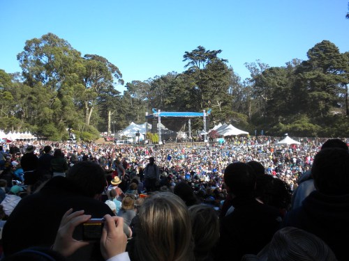 A large crowd gathers at an outdoor event, with a stage and trees in the background under a clear blue sky.