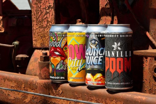 Four colorful beer cans displayed on a rusty metal surface, showcasing various craft beer labels and designs.