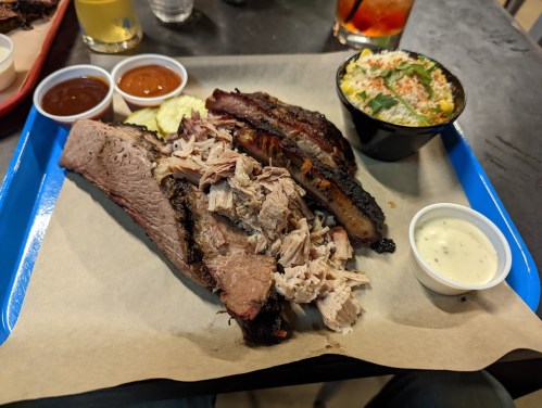 A blue tray with sliced brisket, pulled pork, ribs, sauces, and a side salad on a table.