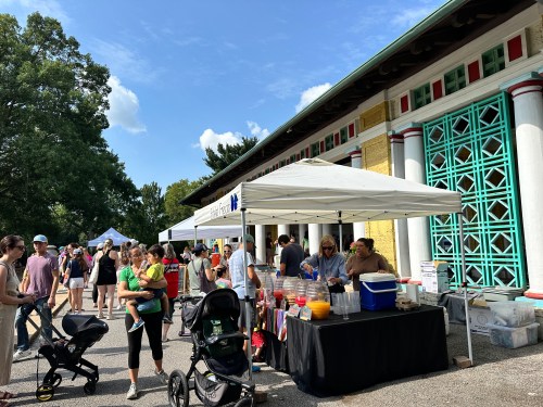 A busy outdoor market scene with people browsing stalls under tents, surrounded by trees and a colorful building.