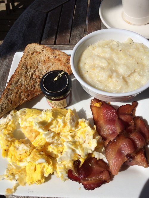 A plate with scrambled eggs, crispy bacon, grits, a slice of toast, and a small jar of jam.